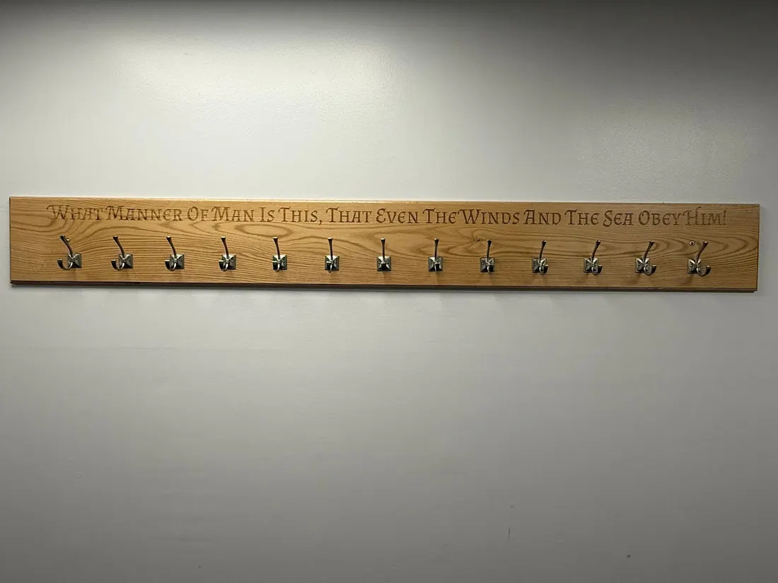 Wall-mounted white oak coat rack built and engraved by Comley Creations, with Matthew 8:27 lettering above a row of metal hooks.